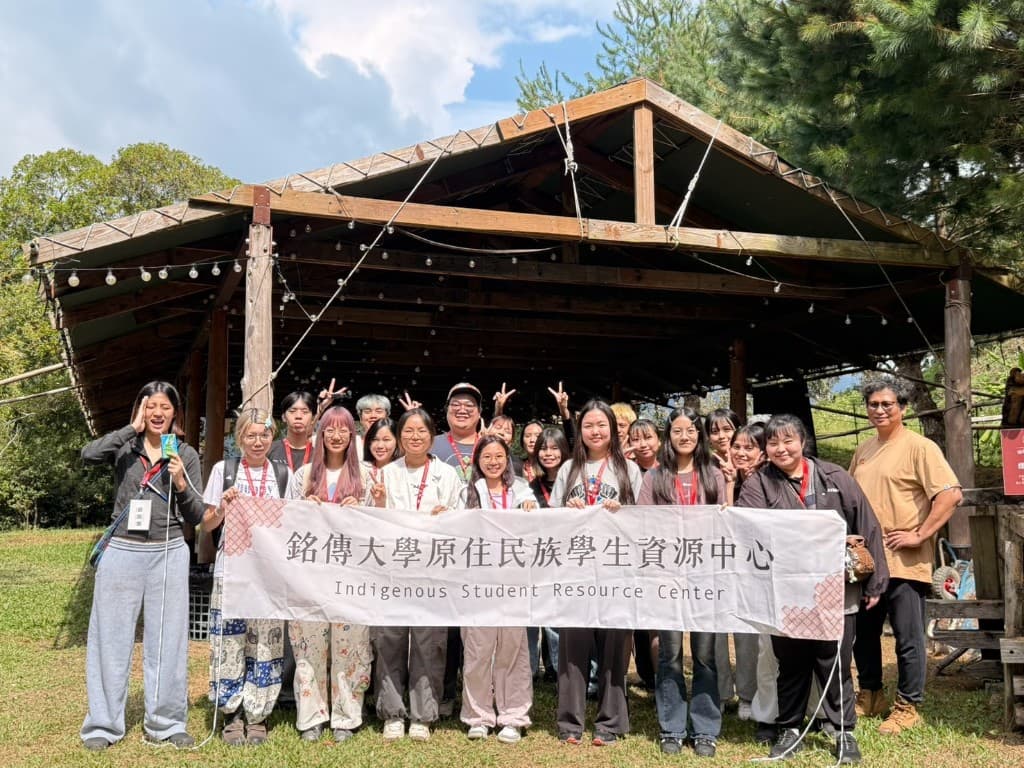 Students and staff of Ming Chuan University’s Indigenous Students’ Resource Center holding the center banner under a wooden pavilion