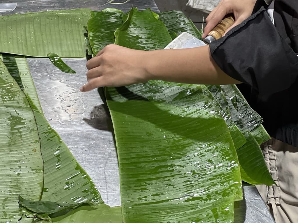 Washed banana leaves being trimmed with a cleaver on a metal table