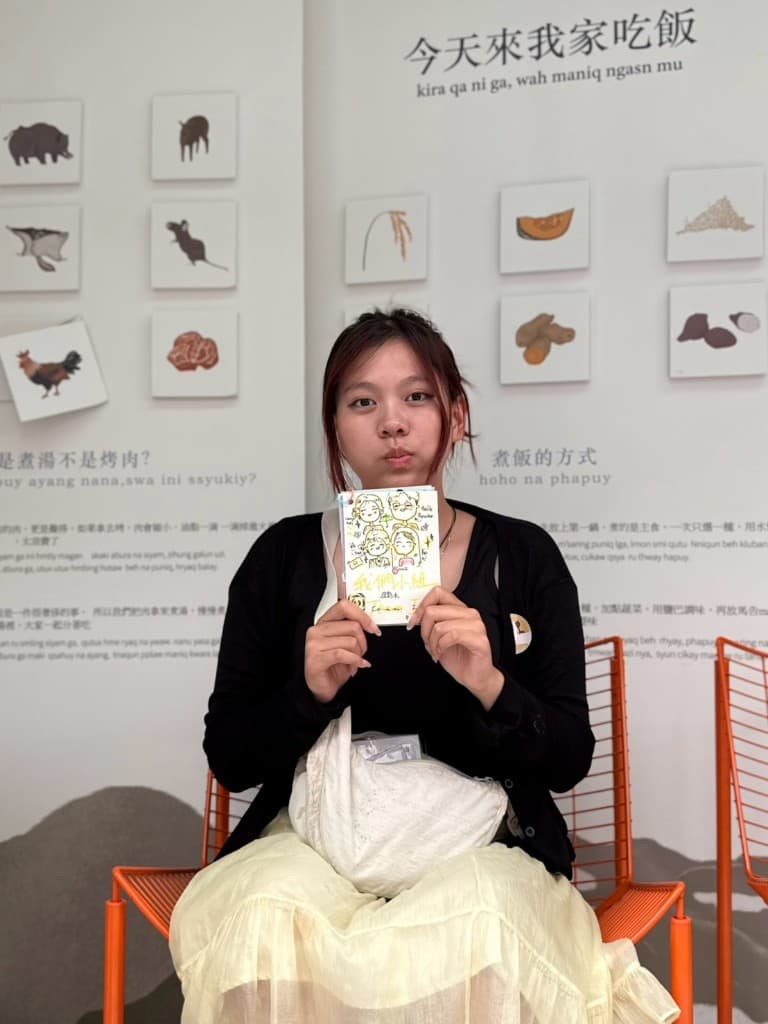 A student seated before the “Come eat at my home today” wall, holding a hand-drawn group card