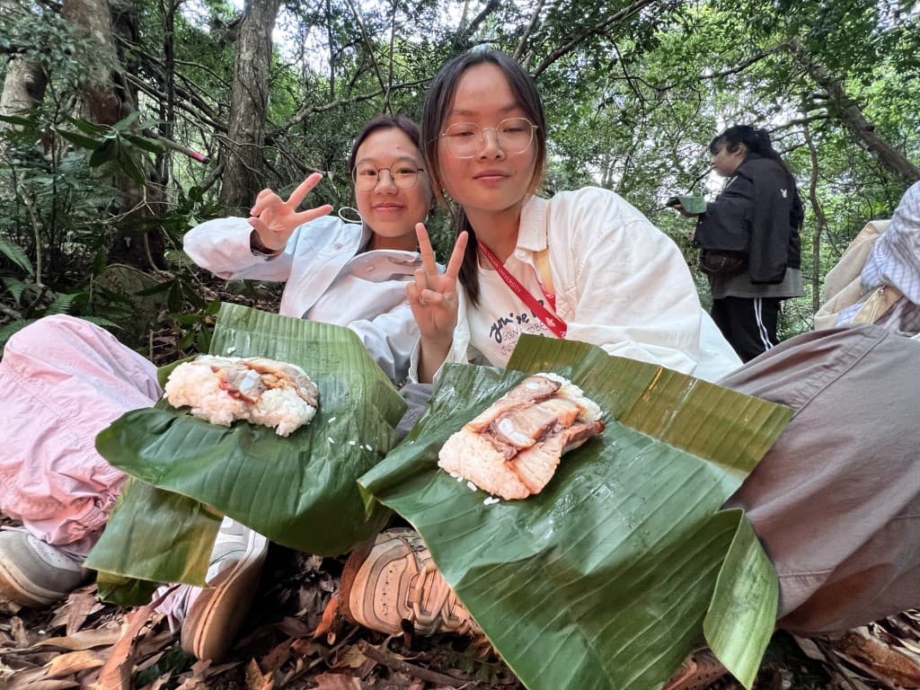 Two students sitting in the forest with banana-leaf meals, making peace signs