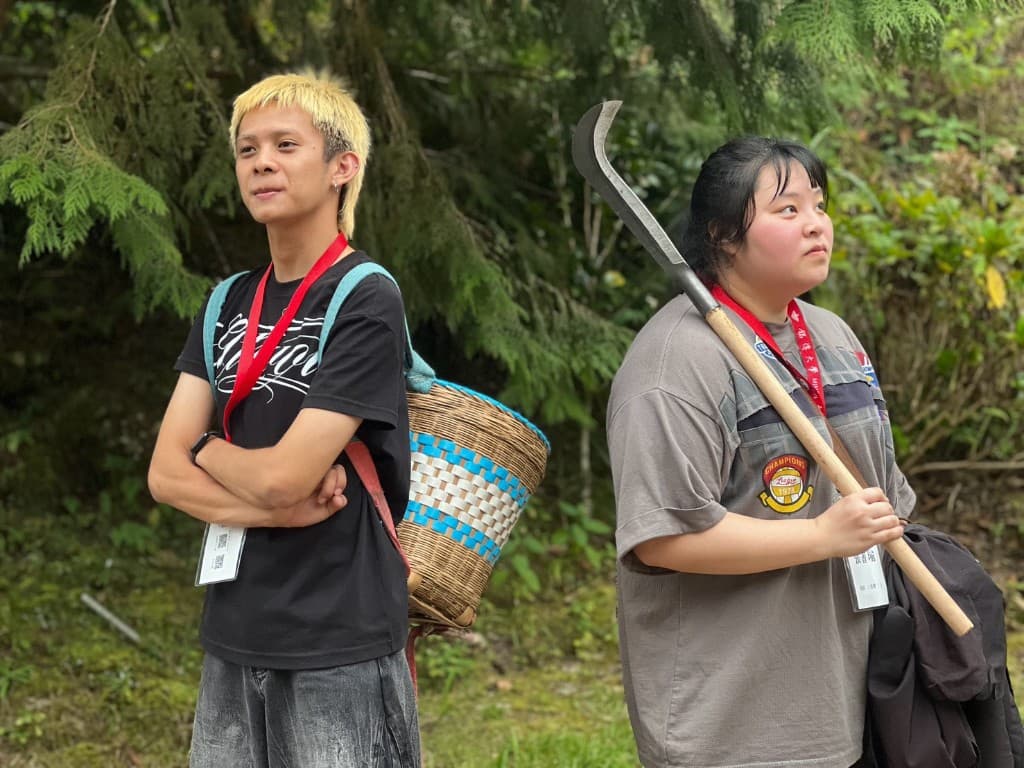 Two students on a forest path, one carrying a woven basket and one holding a sickle